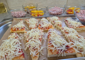 Bezglutenowa pizza przygotowana w kilku wariantach dietetycznych. Na blasze kawałki pizzy z sosem pomidorowym i startym serem, obok miseczki z dodatkami do samodzielnego komponowania porcji przez dzieci.